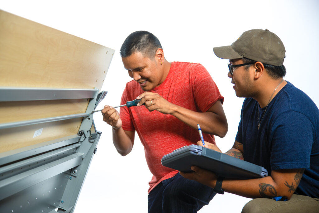 Two people assembling a table, one using a screwdriver while the other takes notes on a clipboard.